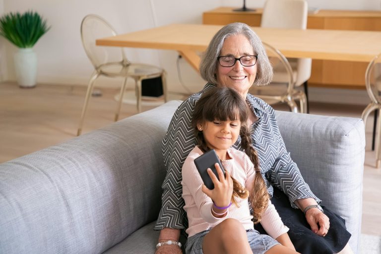 Mujer mayor con su nieta sonriendo y usando un móvil, representando el vínculo emocional y el impacto positivo del precio de audífonos en 2025 en la comunicación familiar.