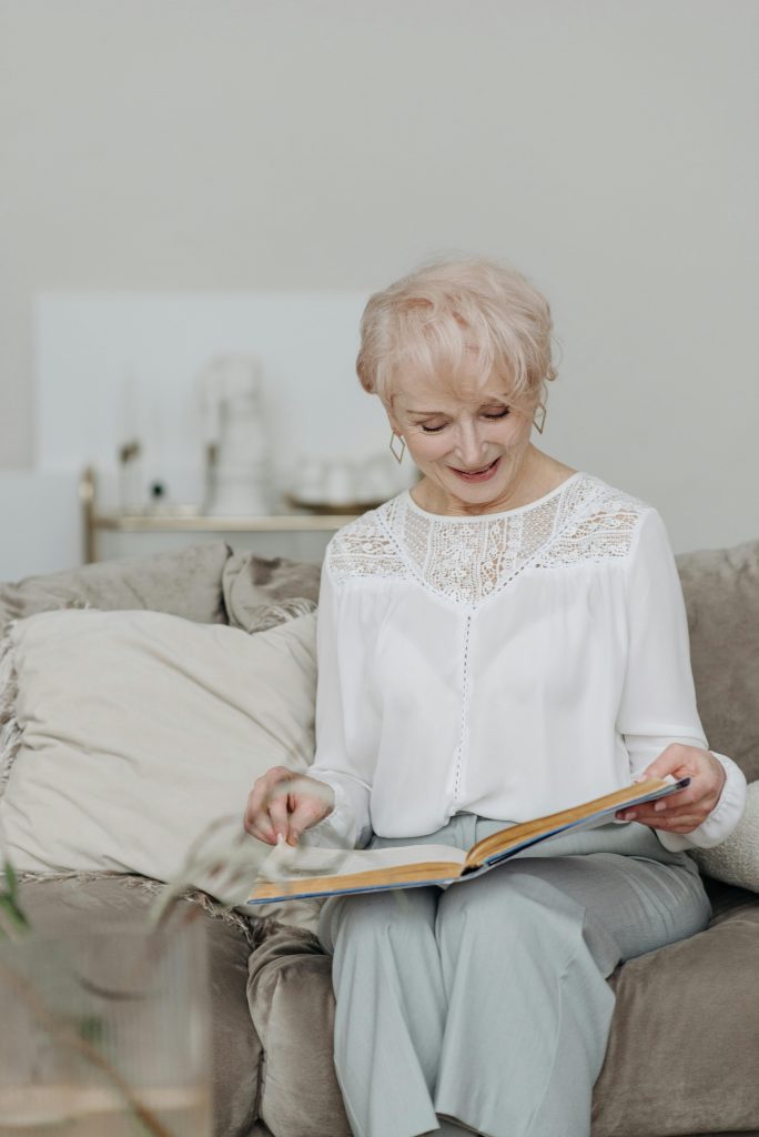 Mujer mayor disfrutando los beneficios audífonos modernos mientras lee un libro en casa.