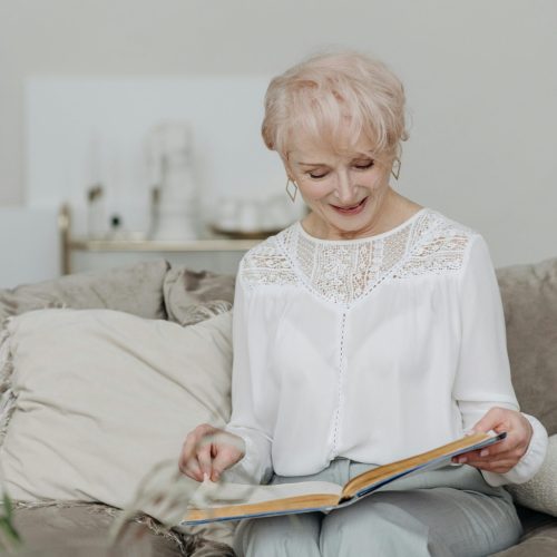 Mujer mayor disfrutando los beneficios audífonos modernos mientras lee un libro en casa.